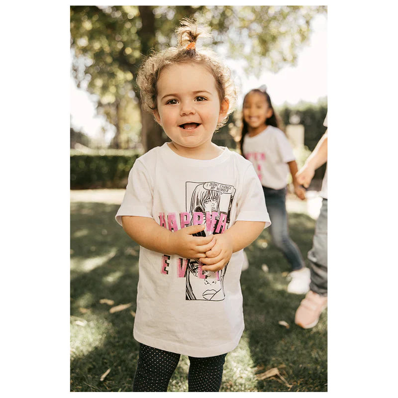 Child standing in a park, wearing a white "Didn't Change My Number" organic youth T-shirt from Billie Eilish merch, smiling at the camera