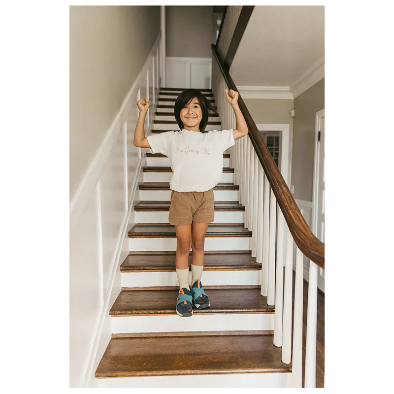 Child standing on stairs wearing a white "I'm Getting Older" organic youth T-shirt from Billie Eilish merch, with arms raised in a joyful pose