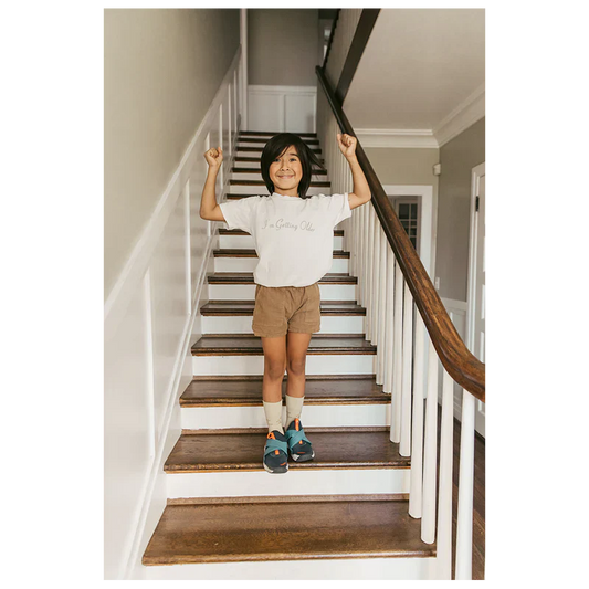 Child standing on stairs wearing a white "I'm Getting Older" organic youth T-shirt from Billie Eilish merch, with arms raised in a joyful pose