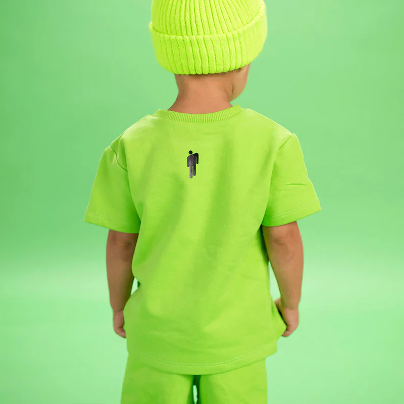 Child wearing a bright green Billie T-Shirt, showing the back with a black stick figure logo, part of the Billie Eilish merch collection