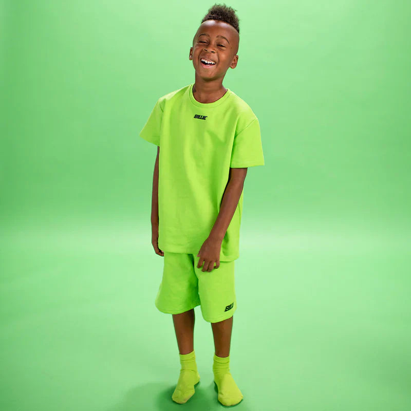 Child standing and smiling while wearing a bright green Billie T-Shirt and shorts from Billie Eilish merch.