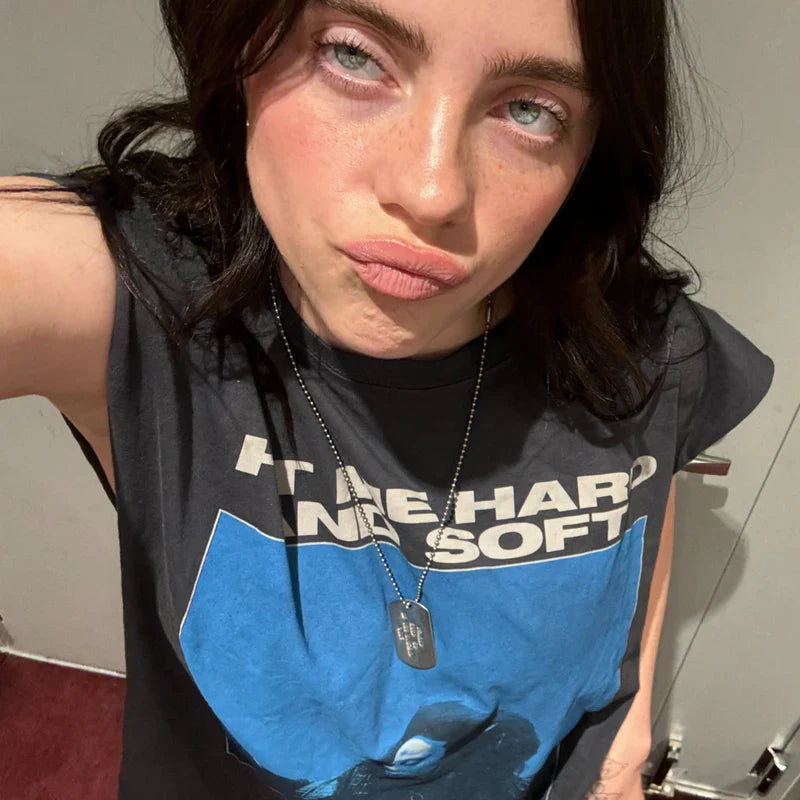 Billie Eilish taking a selfie while wearing the Tracklist Cutoff Muscle T-Shirt showcasing the graphic design and text 'HIT ME HARD AND SOFT