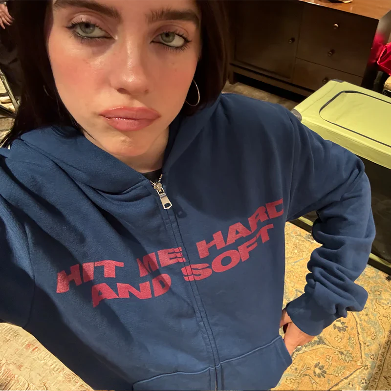 Billie Eilish wearing the Blue & Red Zip Hoodie with 'HIT ME HARD AND SOFT' text in red on a blue hoodie.