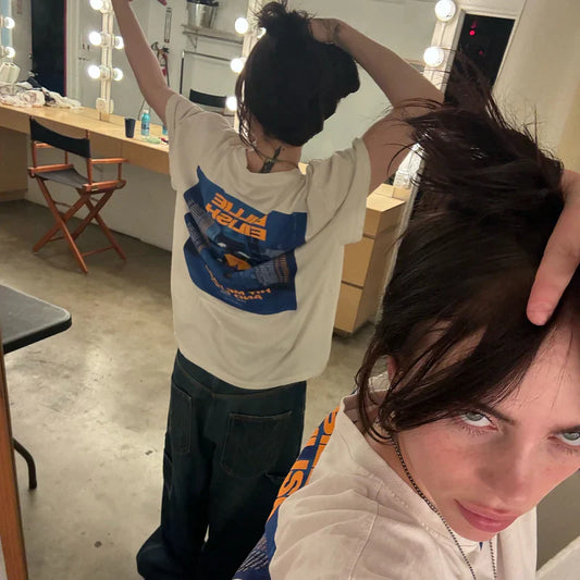 Billie Eilish taking a mirror selfie wearing the Hit Me Hard And Soft Admat Poster Tee, featuring a colorful tour design on the back.