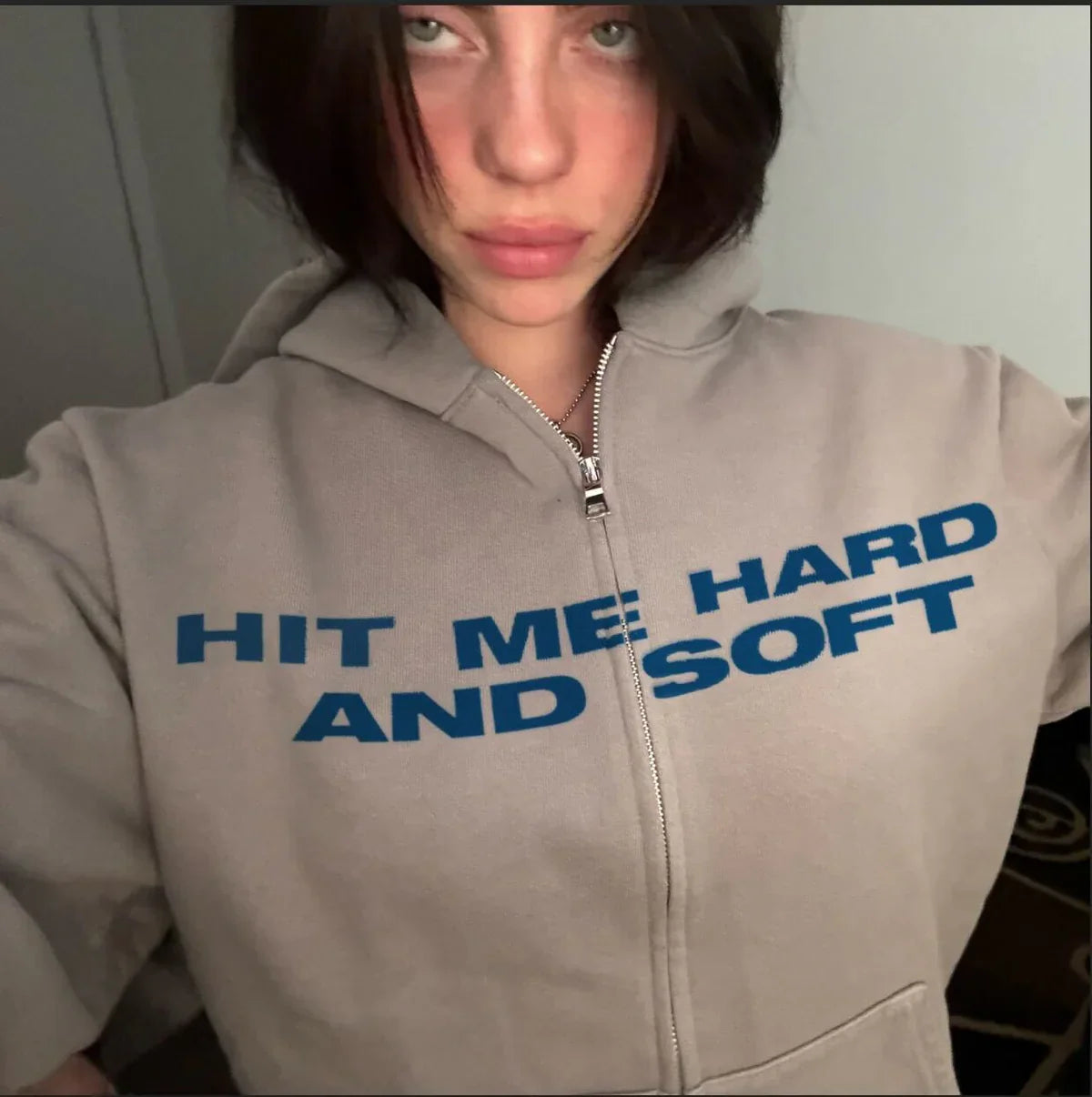 Billie Eilish wearing the Hit Me Hard And Soft Grey Zipper Hoodie with 'HIT ME HARD AND SOFT' text in blue